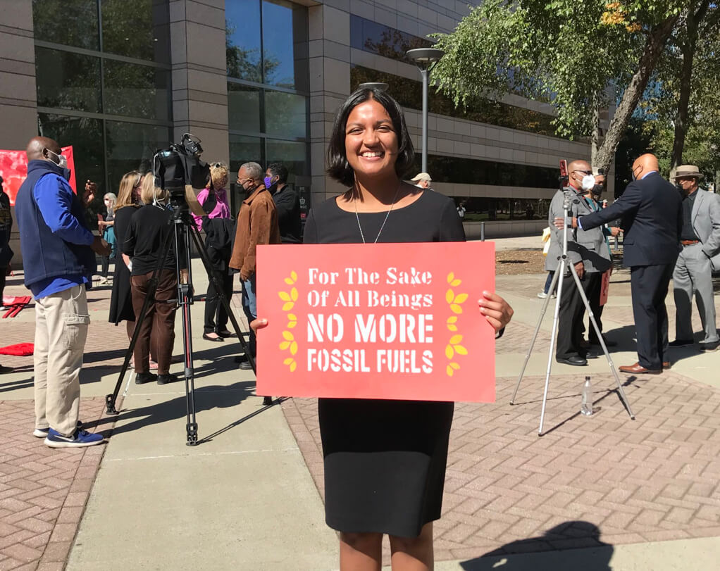 Shruti Agrawal stands with a sign that says "For the sake of all beings, no more fossil fuels" in front of reporters at a rally.