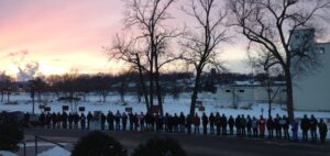 A line of about 30 people standing side by side as a pipeline vigil in Minnesota. The sun in setting behind them and there is snow on the ground.