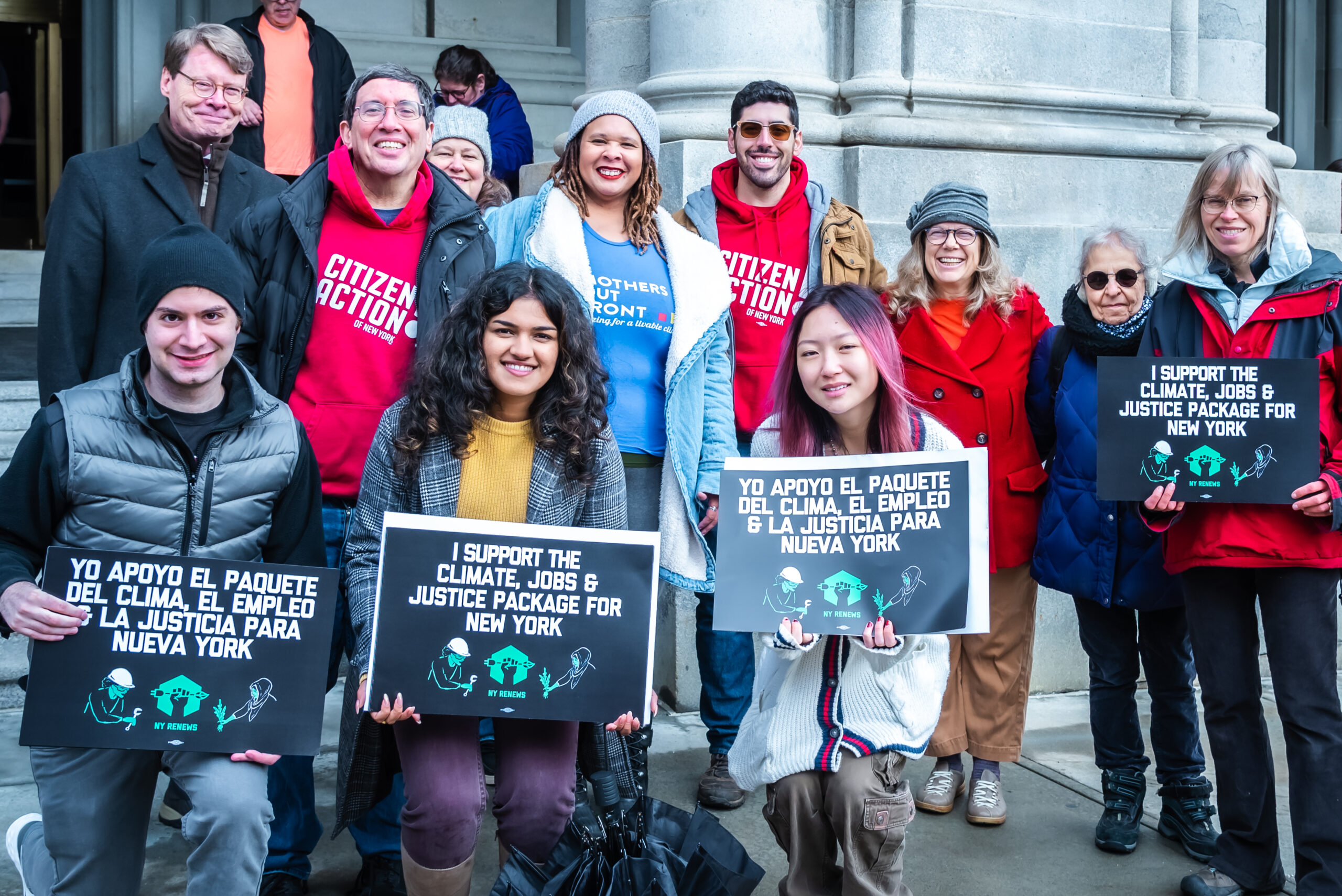 Members of NY Renews standing with climate justice signs at a rally in Albany, NY in support the climate, jobs, and justice package. They are smiling at the camera.